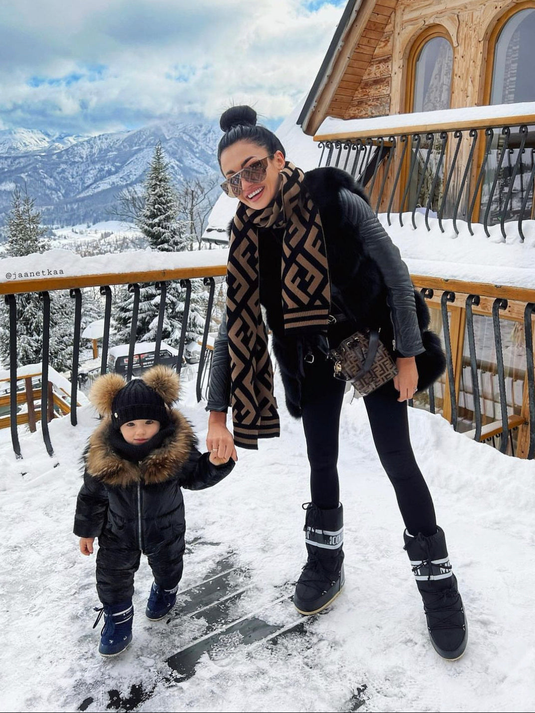 Woman wearing a fox fur vest and child standing on a snowy deck with mountains in the background