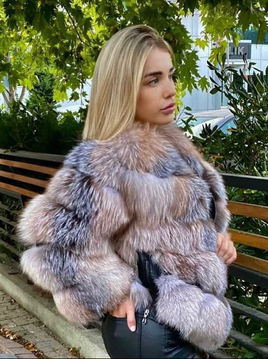 Woman wearing a frost fox fur coat standing outdoors with greenery and a building in the background