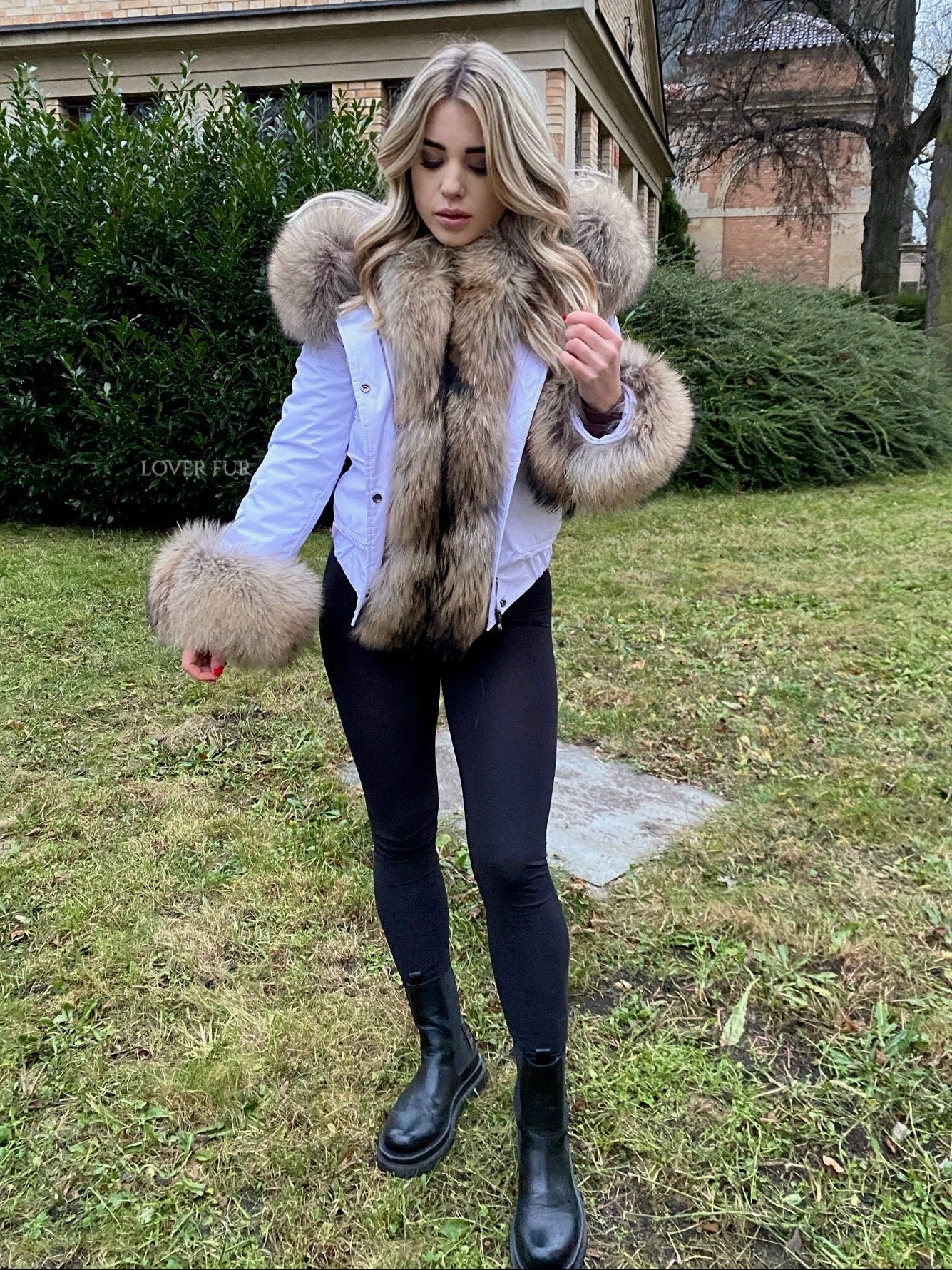 Woman wearing a raccoon fur-trimmed coat in an outdoor setting with a building and trees in the background.