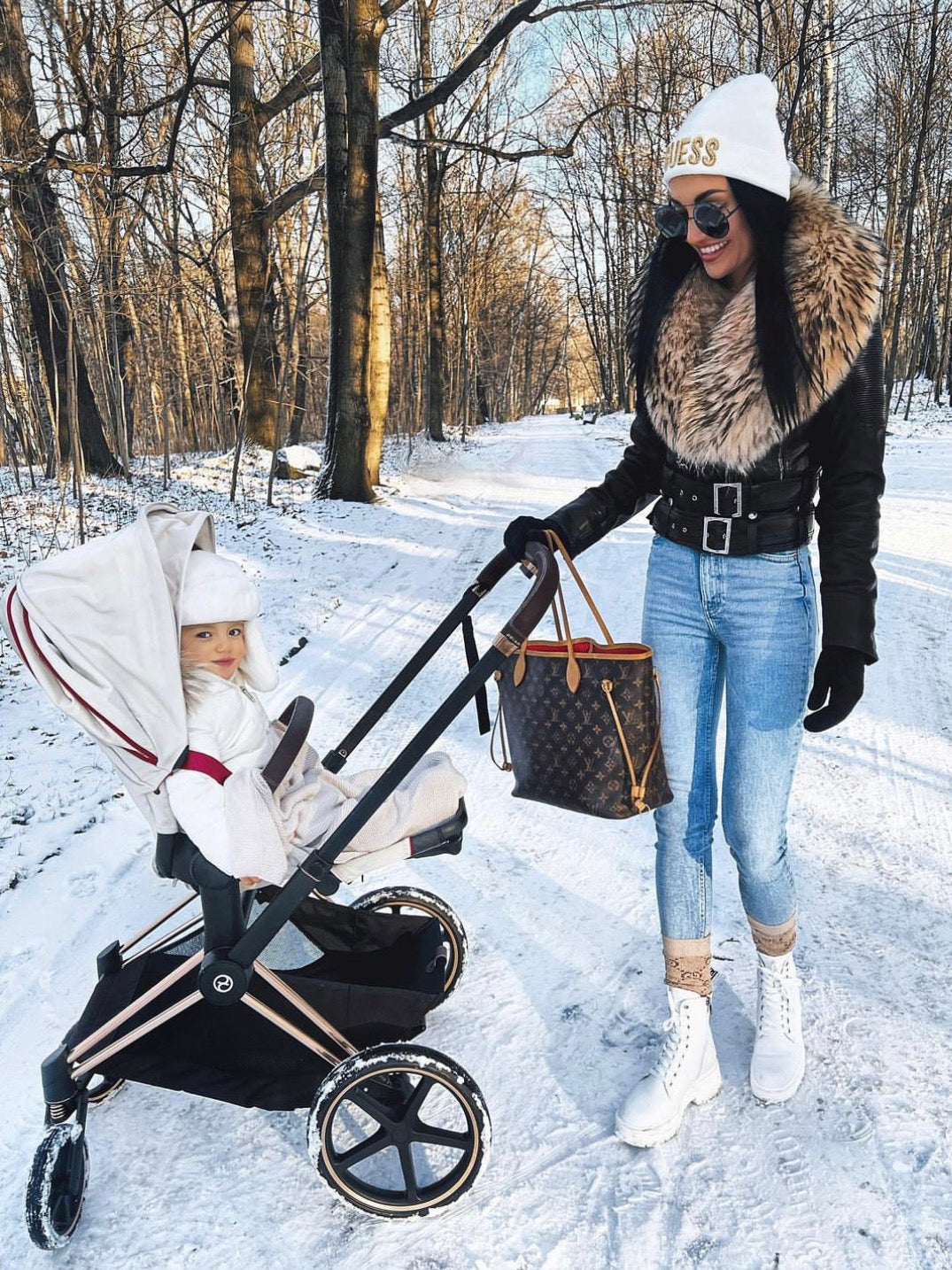 Woman with a leather jacket and raccoon fur pushing a stroller with a child in a snowy forest