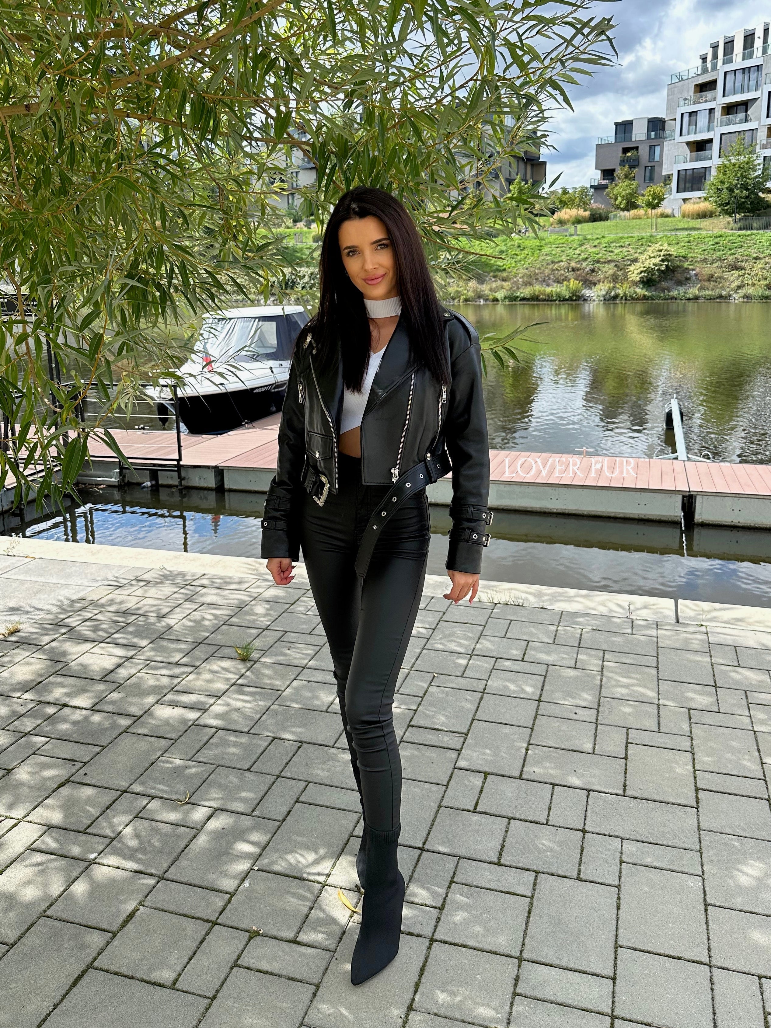 Woman in black leather jacket and pants standing by a waterfront with trees and buildings in the background.