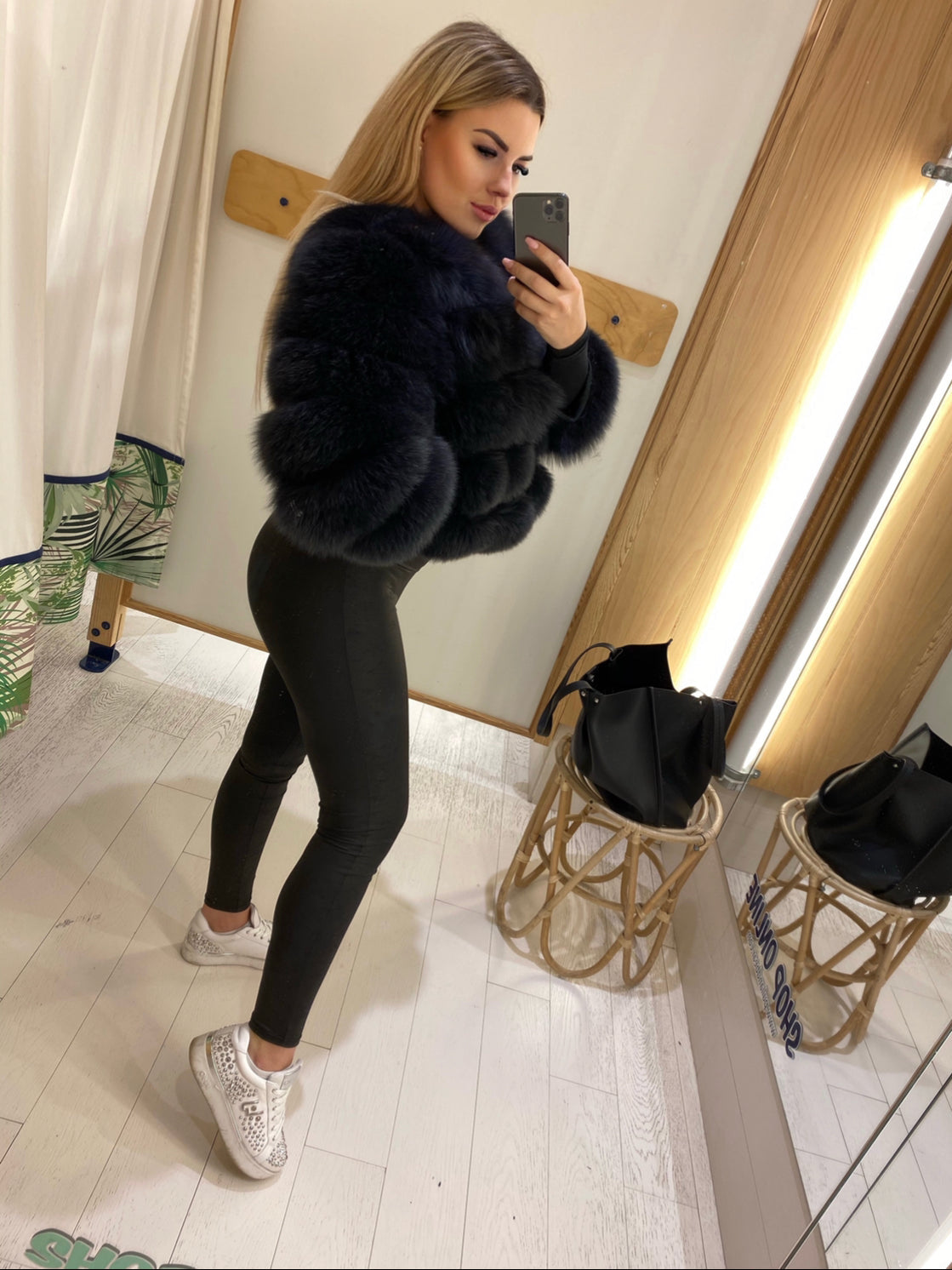Woman taking a mirror selfie wearing a black modular lambskin fur jacket and leggings in a room with wooden floors and a mirror.