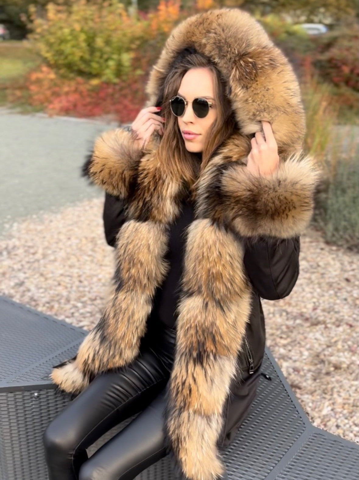 Woman wearing a waterproof fur-lined parker sitting outdoors with autumn foliage in the background