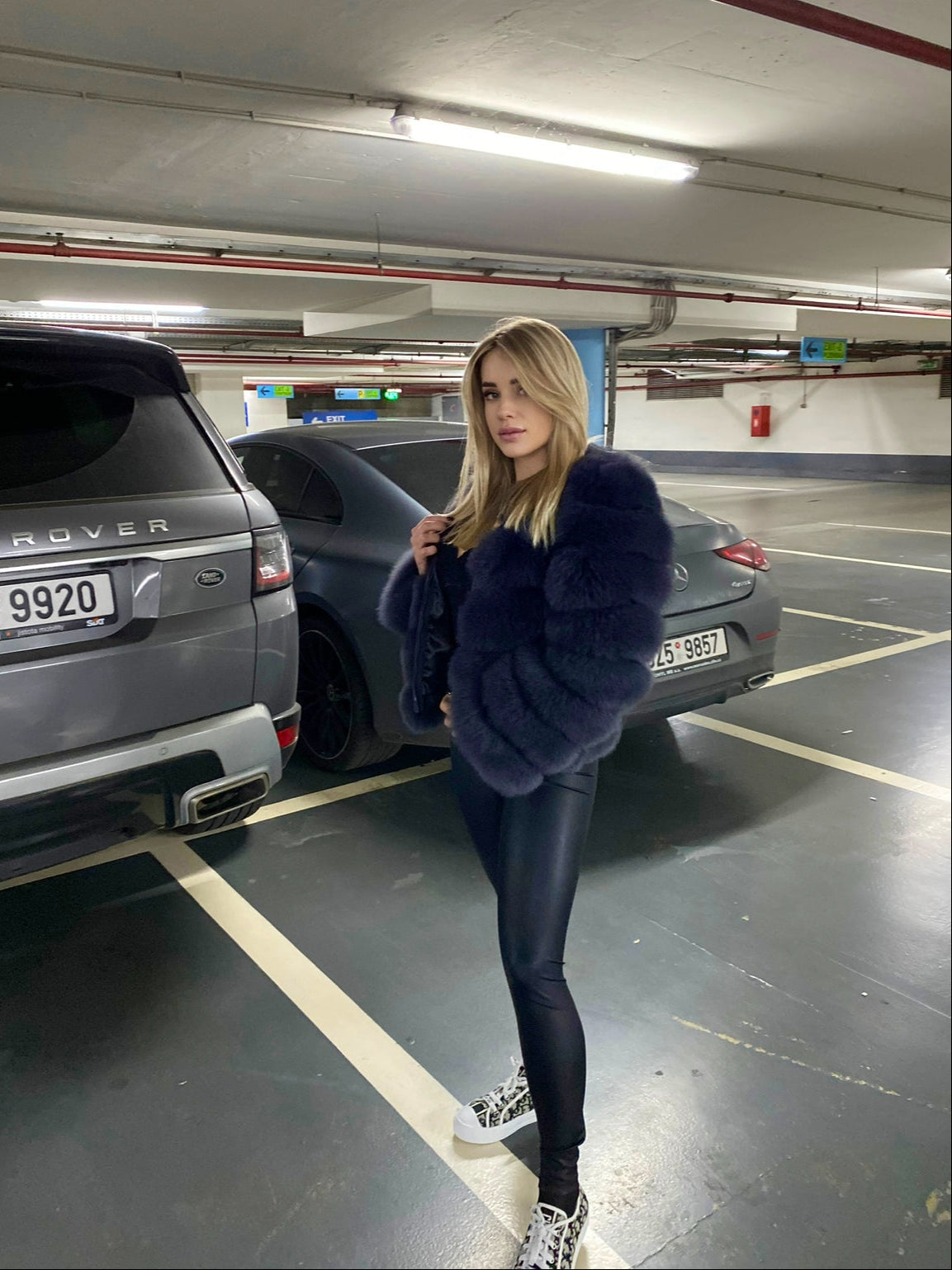 Woman in a blue fox fur coat standing in a parking garage with cars and visible license plates.