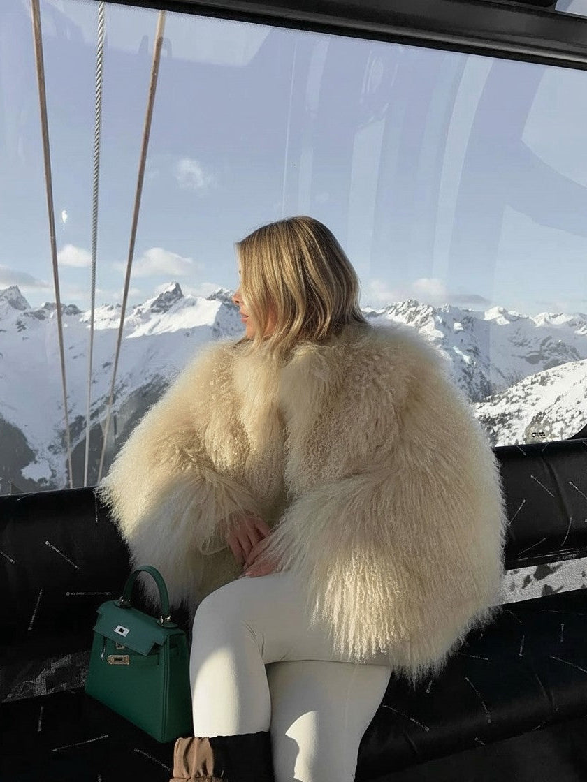 Person wearing a fluffy white coat sitting on a ski lift with snowy mountains in the background