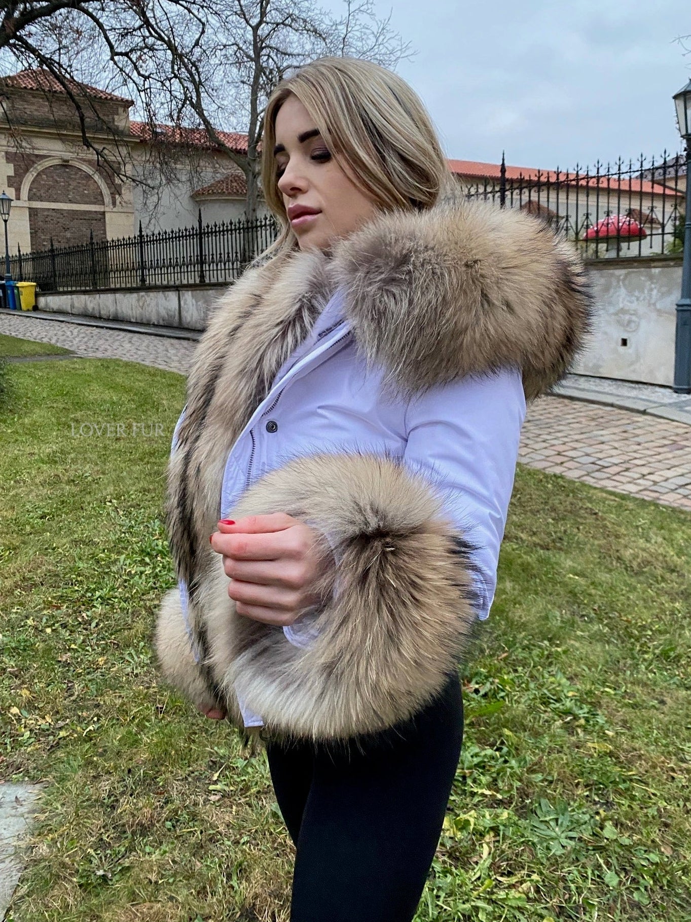 Woman wearing a raccoon fur vest outdoors with a building and trees in the background