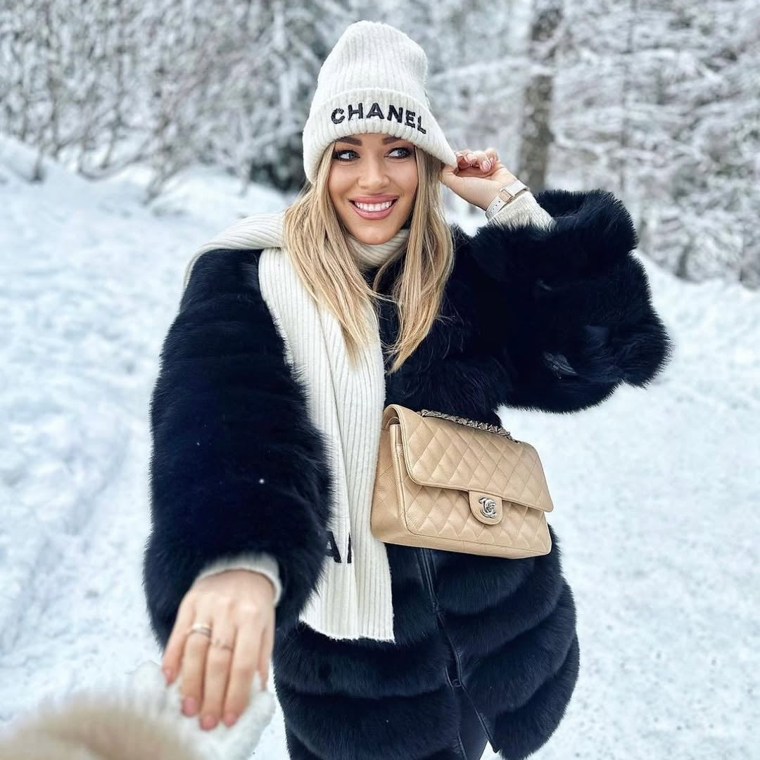 Woman in a snowy landscape wearing a Chanel beanie and coat, holding a matching handbag.